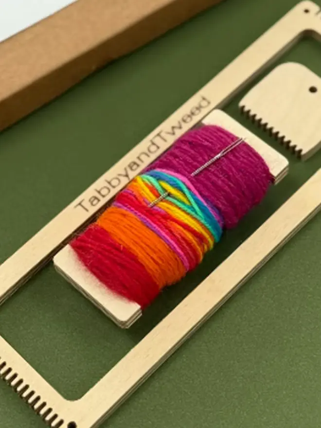 Rectangular beige loom with bright rainbow coloured yarns on a spool and wooden comb.
