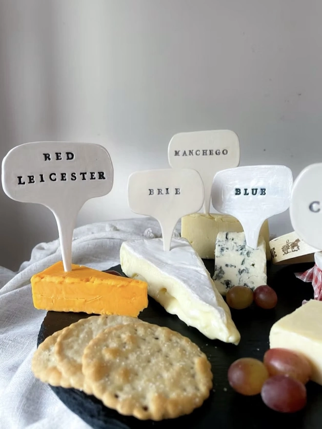 Ceramic Cheese Markers, a group of ceramic cheese markers embedded in a cheese platter. They read: 'Red Leicester, Brie, Manchego, Blue'. 