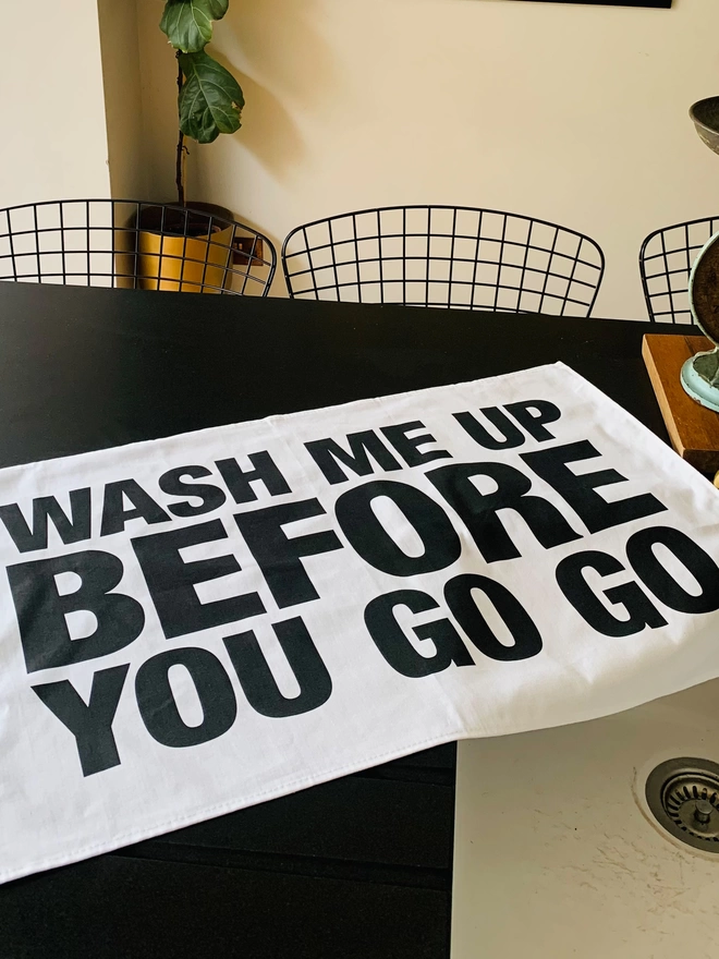 London Drying Wash Me Up Before You Go Go black screen printed text on white tea towel laying on black kitchen counter