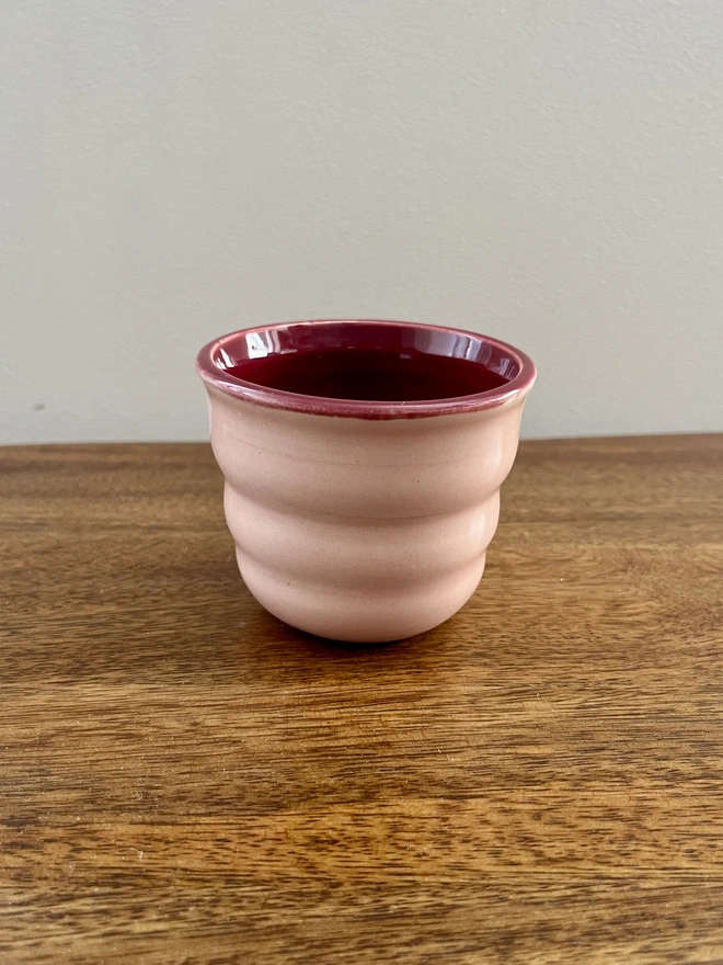 mini ceramic tumbler with wavy sides on a wooden table. the tumbler has a blush pink glaze on the outside and sangria burgundy on the inside and rim. 