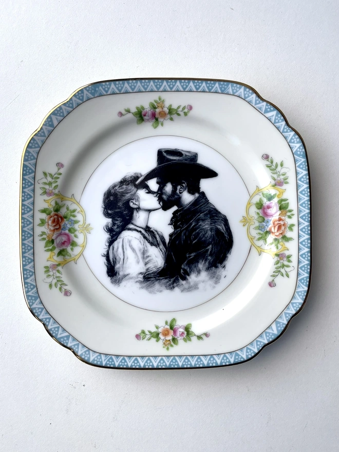 a vintage plate with an ornate border has a black and white vintage illustration of a cowboy and cowgirl sharing a kiss in the centre