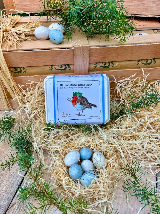 Delicious christmas robin eggs nestled in a wood wool nest in front of a Chocolate Detective egg box surrounded by wood wool and fresh juniper branches.