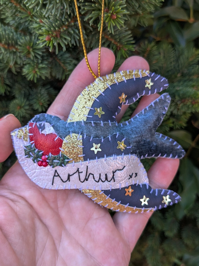 Hand stitched personalised swallow Christmas decoration made from vegan-friendly faux leather and velvet, featuring gold stars, festive holly and a cream banner personalised with a name, hanging from a gold cord on a Christmas tree.