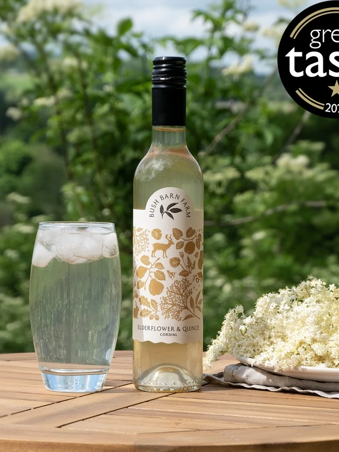 Elderflower & Quince Cordial (Set Of 1, 2, 3 Or 4), a glass of elderflower sitting next to a bottle of elderflower on a wooden table. 