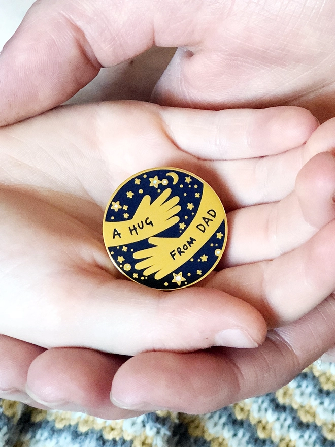 A young child holding a navy and gold a hug from dad pin badge in their hands