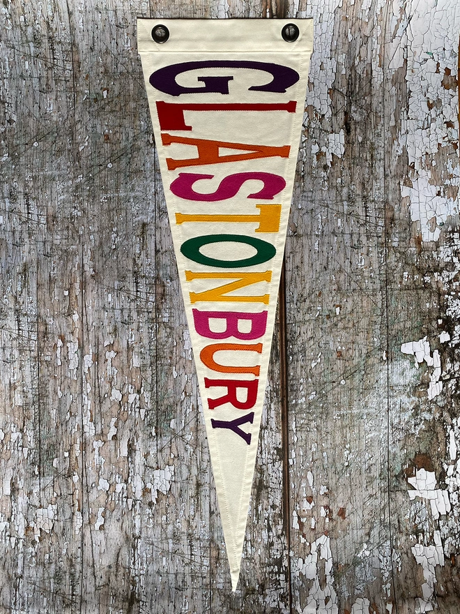 An ivory canvas pennant flag hung vertically on a wooden wall with the word Glastonbury written in purple, red, orange, pink, yellow and green canvas letters