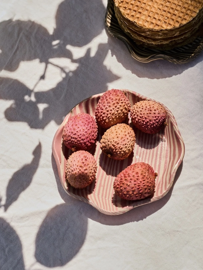 Striped Trinket Dish, a colourful striped trinket dish sitting on a grey surface. It is accompanied by some lychees. 