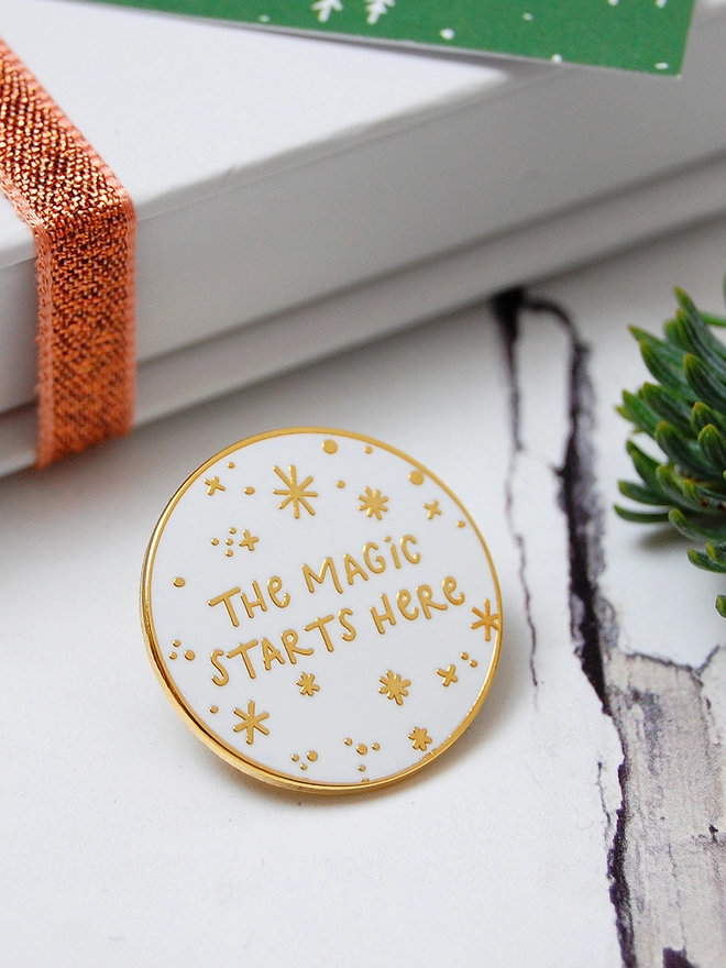 A white and gold enamel pin badge with a starry design and the words "The magic starts here" is beside a gift box on a white wooden table.