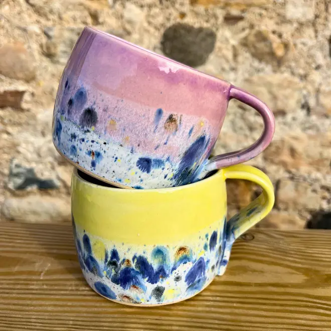 Shoreline Large Round Mug, a pair of colourful mugs stacked on top of each other on a wooden surface against a brick backdrop. 
