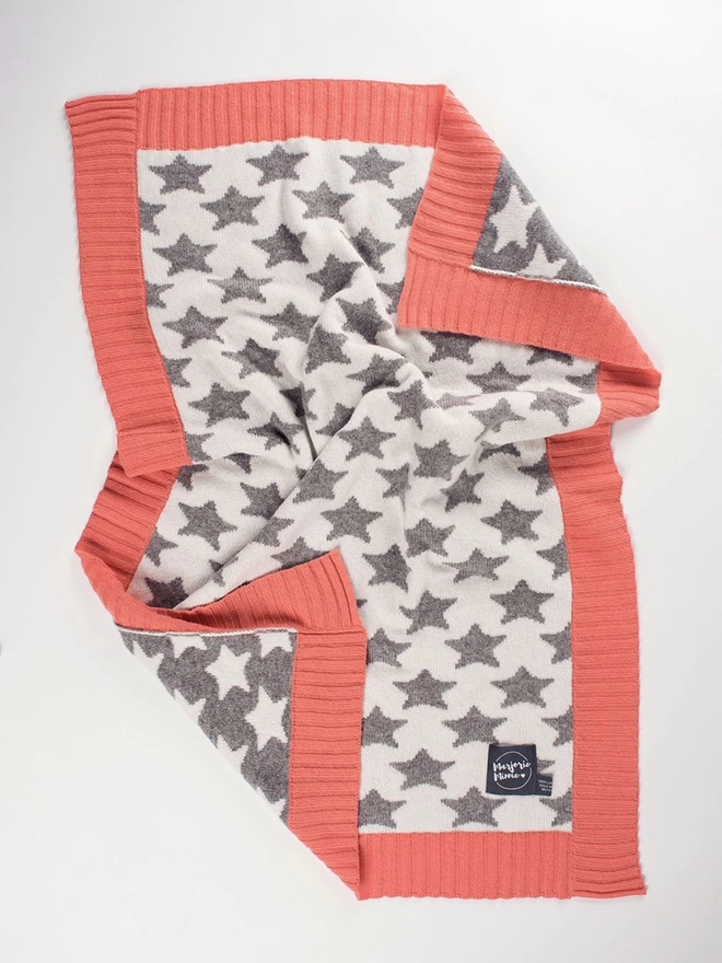A flatlay of a knitted baby blanket with grey and white star pattern and coral pink ribbed trim. The corners are slightly ruffled and turned back to reveal the reverse colourway.