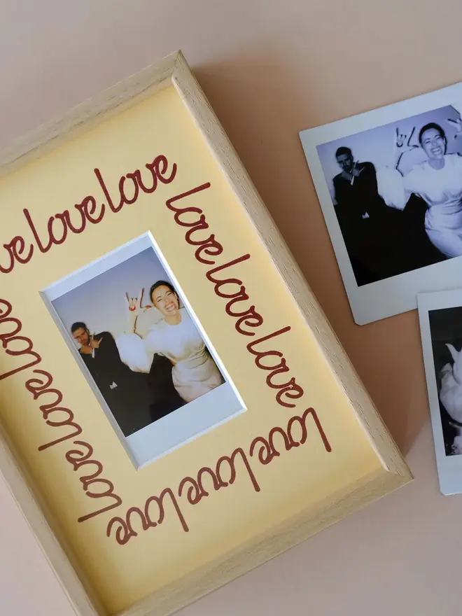'Love' instax mini mount frame with a yellow mount featuring a decorative frill border around a centered Instax Square photo, displayed on a neutral beige wall.