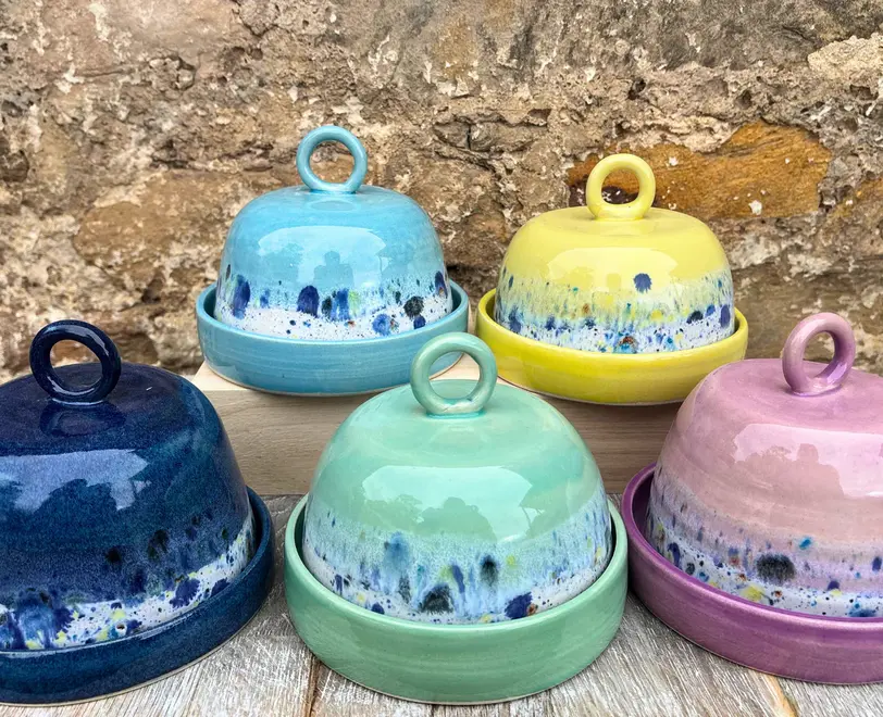 Shoreline Butter Dish, a collection of colourful butter dishes placed on a wooden surface against a brick backdrop. 