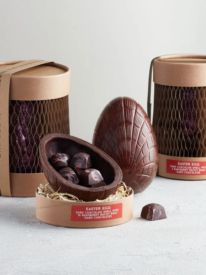 Dark Chocolate Easter Egg With Raspberry Rocky Road, a chocolate egg sitting on a white surface. It is accompanied by another chocolate egg that is opened showing the chocolates within. 