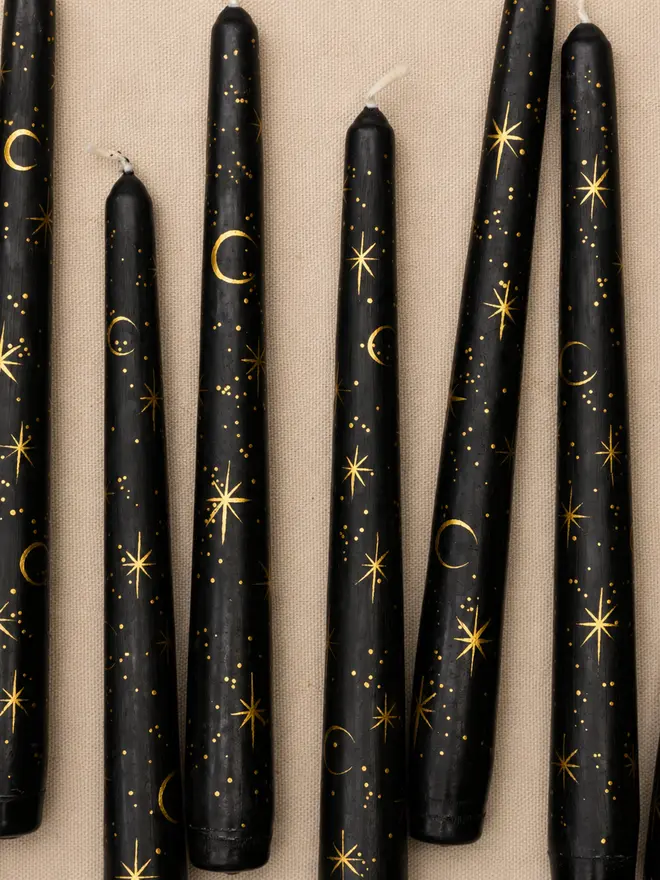 Close-up of hand-painted black taper candles featuring delicate gold moon and star illustrations on a natural linen background.