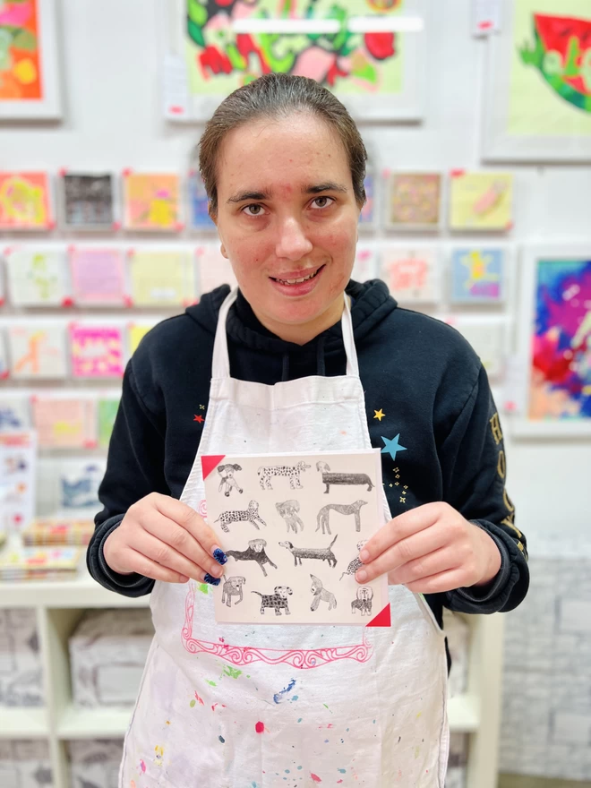 Artist holding a card featuring hand drawn black dogs arranged in a pattern on a white background