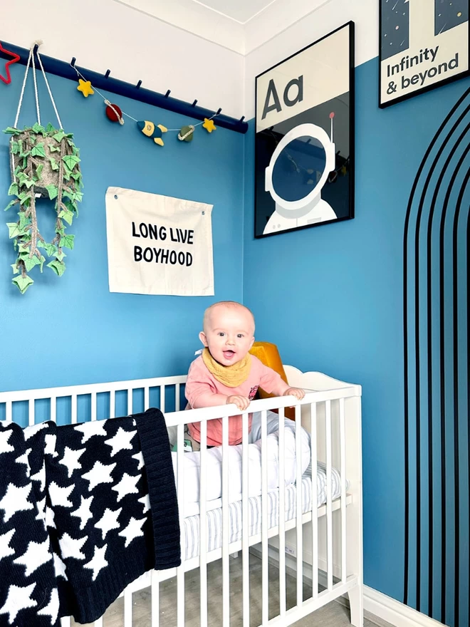 A view of a modern space themed nursery painted a vibrant blue. A baby stands in his cot, holding onto the rail laughing. A black and white monochrome star blanket is draped over the side. A monochrome print of  ‘A is for Astronaut’ hangs on the wall.