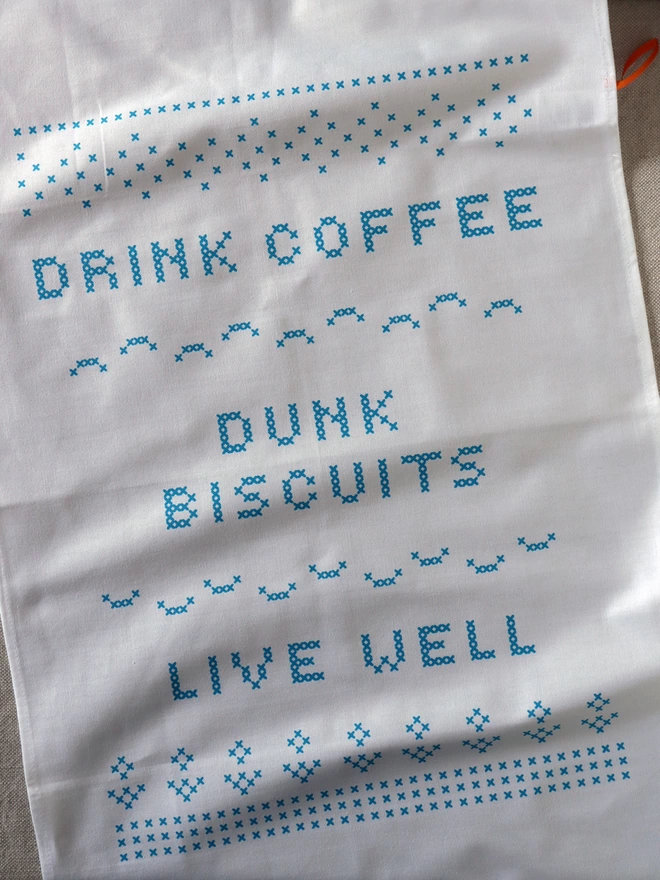 A Drink Coffee, Dunk biscuits, live well cross-stitch style printed white cotton tea towel laid flat on a table