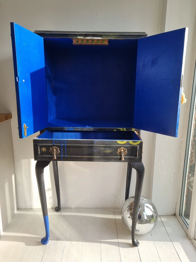 Elegant cocktail cabinet in black with colourful florals and text in chartreuse green that reads 'hot tramp, I love you so', a David Bowie lyric. Heavily distressed to age it. The interior is cobalt blue.