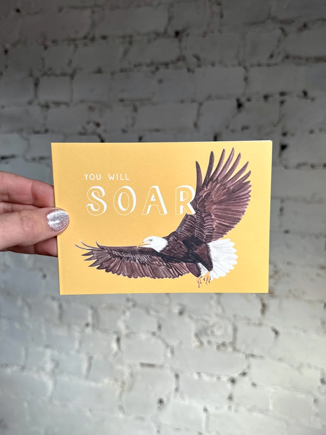 a bright yellow greetings card featuring an illustration of a bald eagle with the text “you will soar”