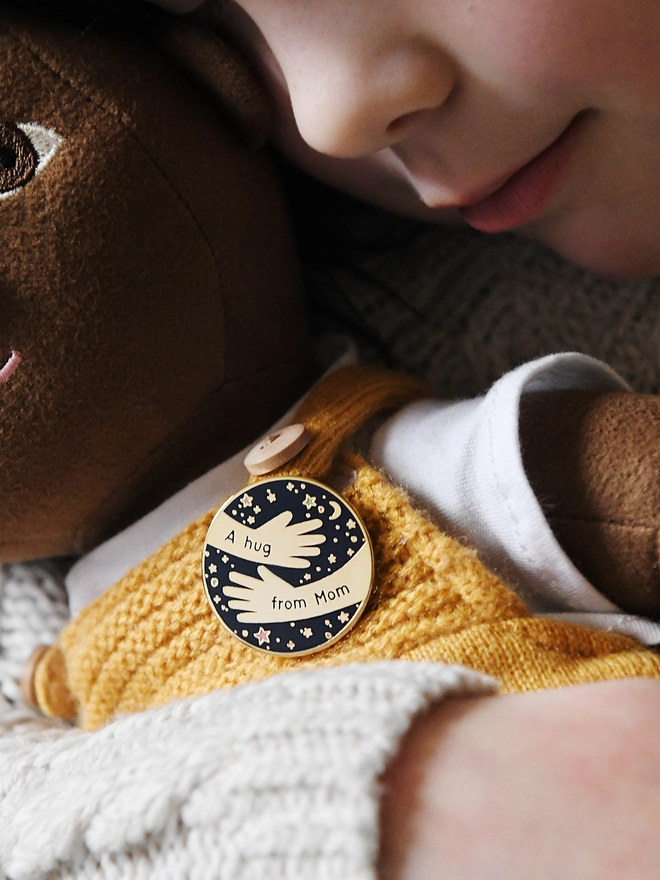 A navy blue and gold enamel pin badge with a hugging arms design and the words "A hug from Mom" is pinned to the clothes of a doll being hugged by a young child.