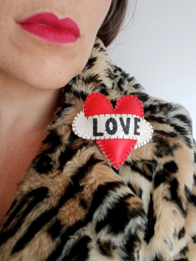 Red Heart Brooch with a white scroll with LOVE stitched on in black lettering, worn on a leopard fur coat. 