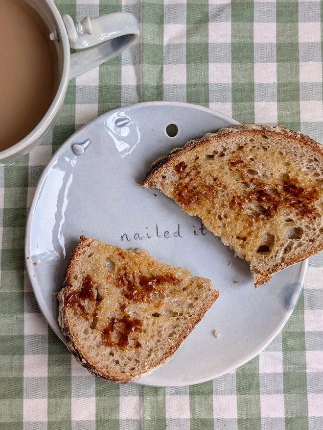 nailed it grey ceramic handmade plate with pun and toast on and pottery nail on a green gingham cloth next to a grey screw tea cup