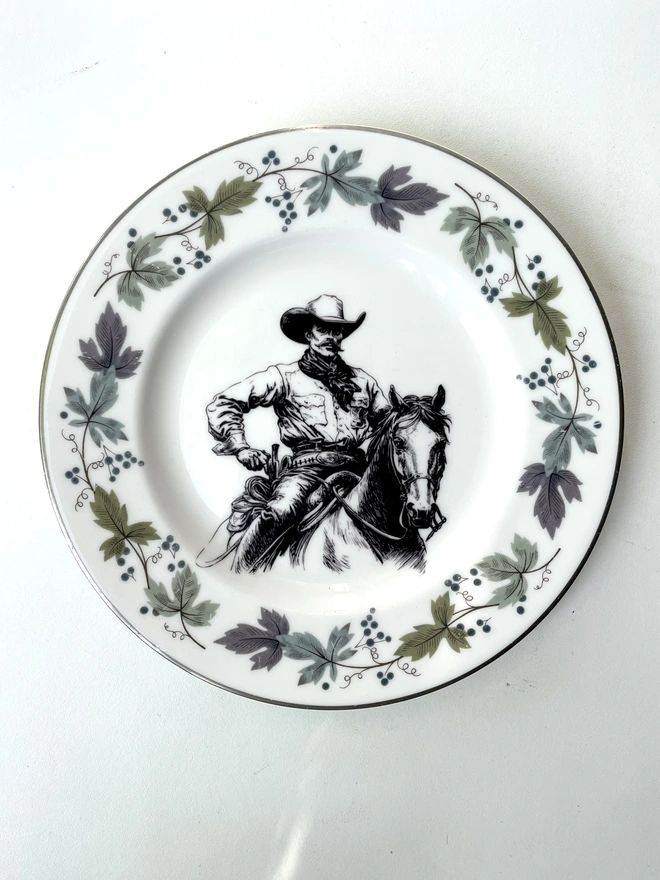 vintage plate hanging on a white wall, it has a decorative gold edge and floral border, with a printed vintage illustration of a cowboy in the middle 