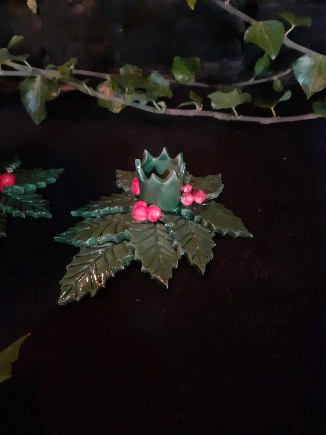 Holly candle holder on a dark surface, showing textured green leaves and three clusters of red berries around the centre.