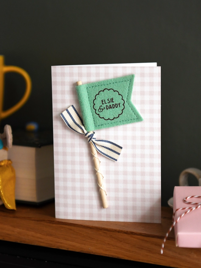 Personalised Father’s Day card with green felt flag reading “Elsie & Daddy”, handmade keepsake card with ribbon and wooden stick detail. The card stands on a wooden mantle beside a book, mug, candle, and wrapped gift.