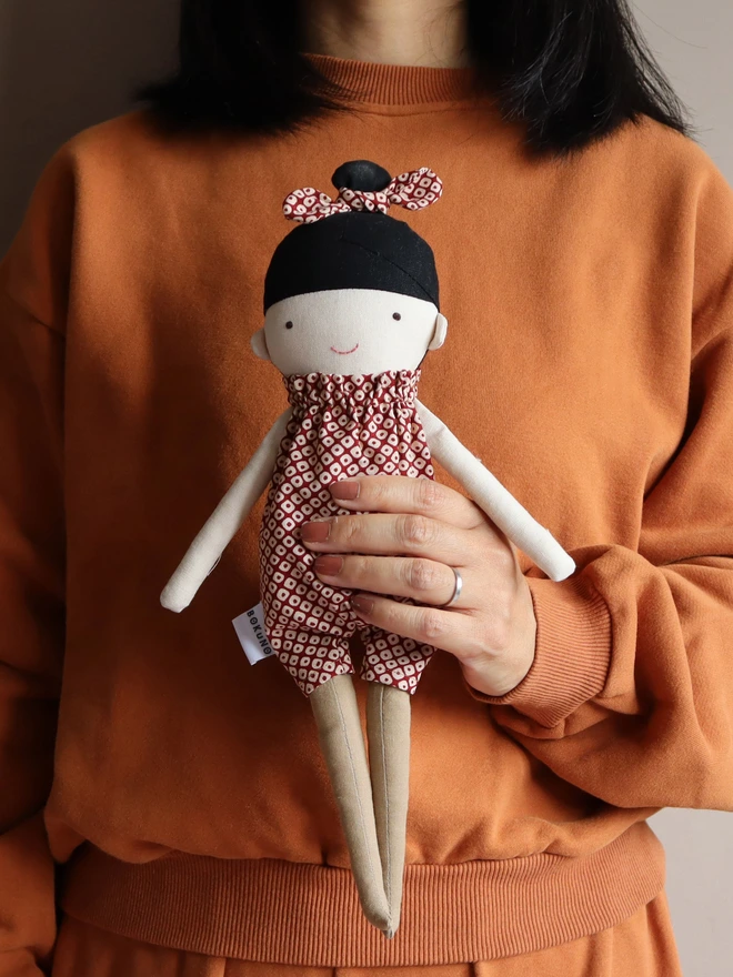 A handmade doll wearing a red and white Japanese shibori patterned jumpsuit with a matching bow in her black hair. The doll is being held by a person's hand.