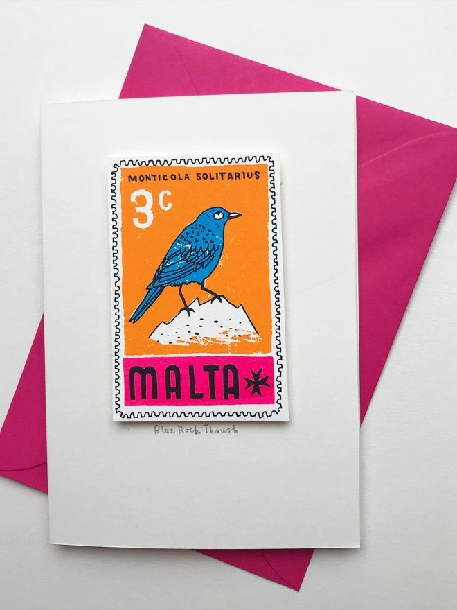Bird Stamp Card Malta/ Blue Rock Thrush, a white card with a colourful bird design in the centre on top of a pink envelope on a white backdrop. 