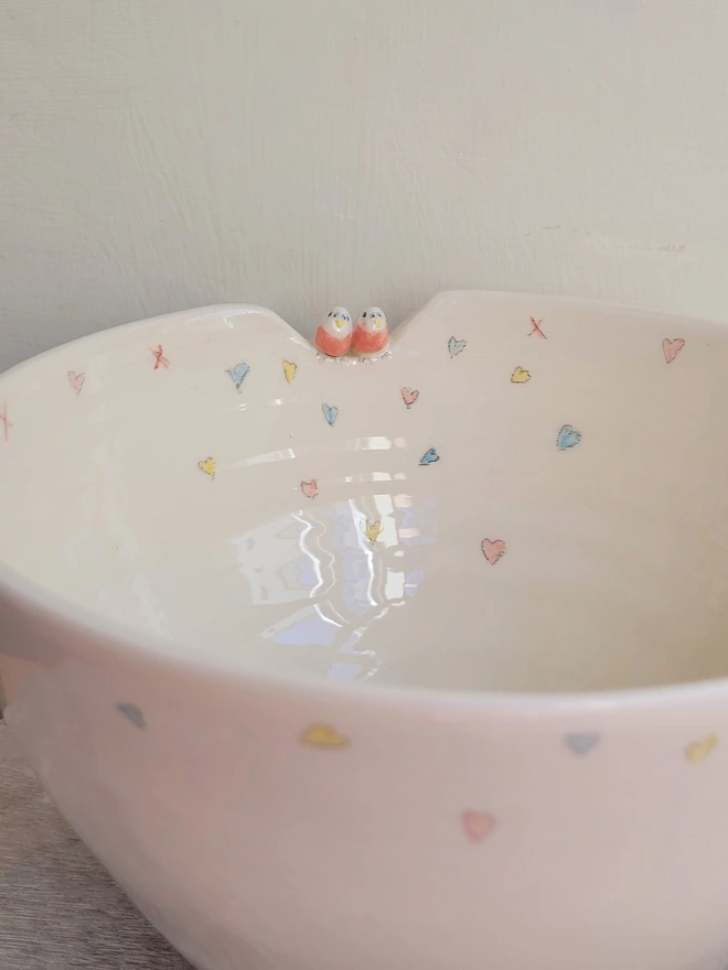 a pottery off white bowl with 2 pink budgerigar ceramic birds perched on the rim 