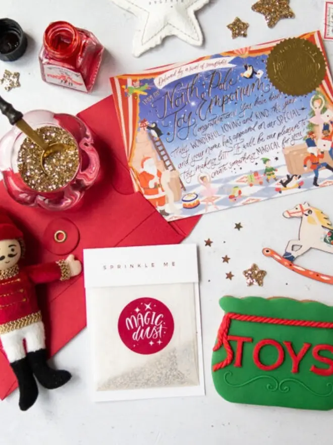 A "Sprinkle me" Magic Dust glassine bag of glitter in front of the illustrated North Pole notecard and a red envelope. Around them are a bowl of gold glitter with a spoon, a felt nutcracker, a rocking horse decoration and an iced "Toys" sack biscuit.