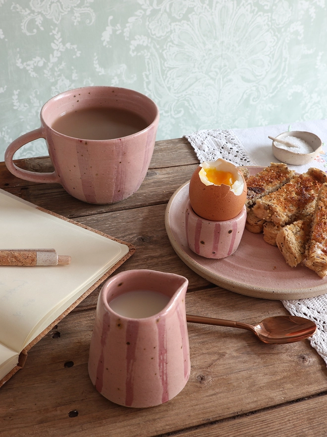 A pink-striped ceramic egg cup sits on a plate with buttered toast, alongside a brass spoon, a notebook, and a matching mug and milk jug on a wooden table.