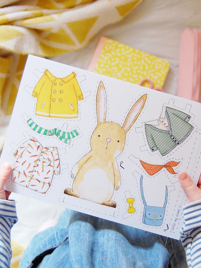 A child is holding a greetings card with an illustrated rabbit paper doll and several outfits on.