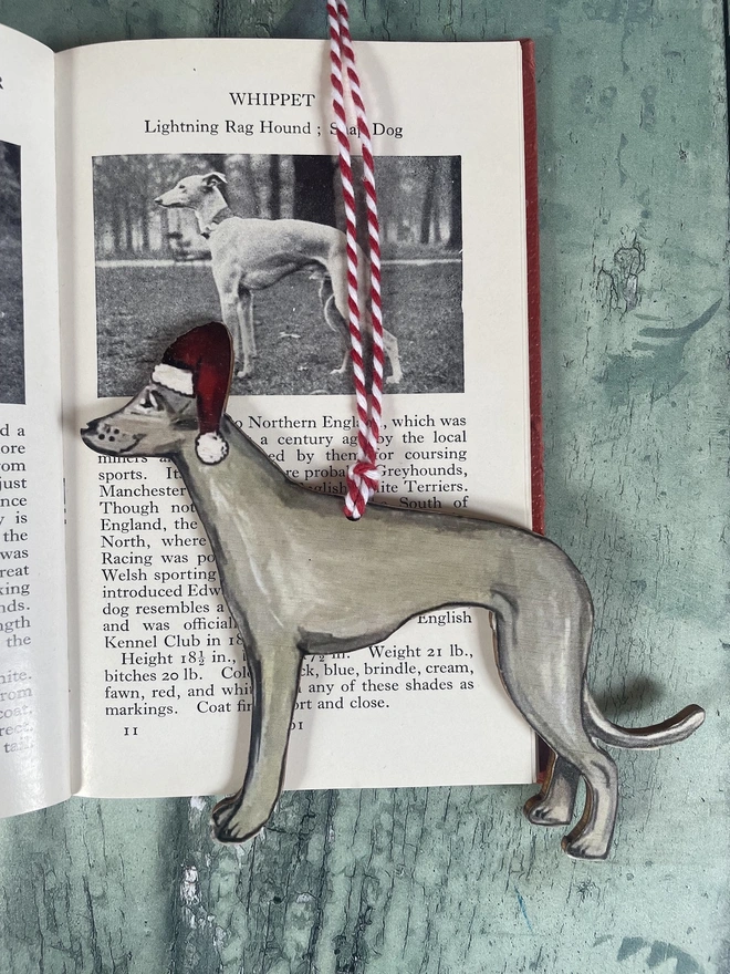 Whippet wooden Christmas decoration wearing a santa hat hung with red and white twine placed onto a printed book about dogs