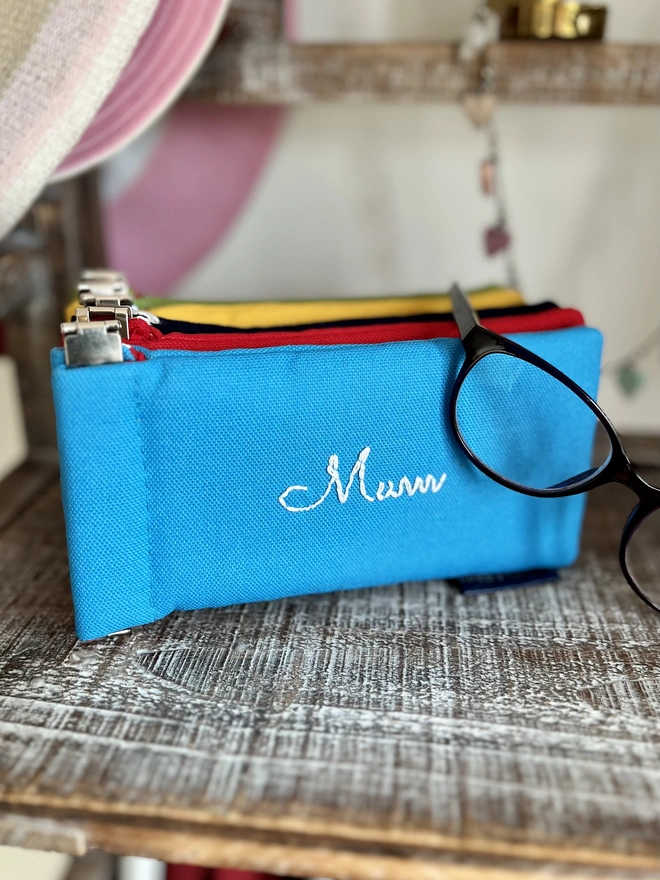 Bright blue soft cotton glasses case with 'Mum' embroidered on it. Spectacles are draped over it and a stack of glasses pouches in different colours behind.