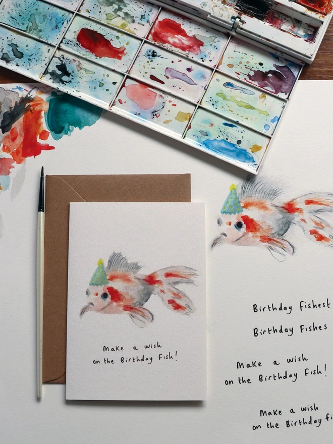 Desk Shot Of The Goldfish Greeting Card Sitting Along Side The Original Hand Painted Watercolour Illustration, Paintbrush and Palette