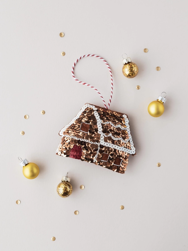 A sequinned gingerbread house ornament on a cream table surrounded by mini baubles