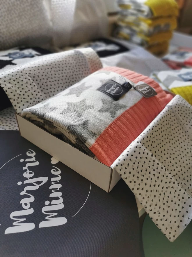  A neatly folded star blanket is shown packed inside a box and wrapped with tissue paper. The box is sitting on a table surrounded by other gift wrapped products. Beneath the box is a grey Marjorie Minnie branded gift bag.