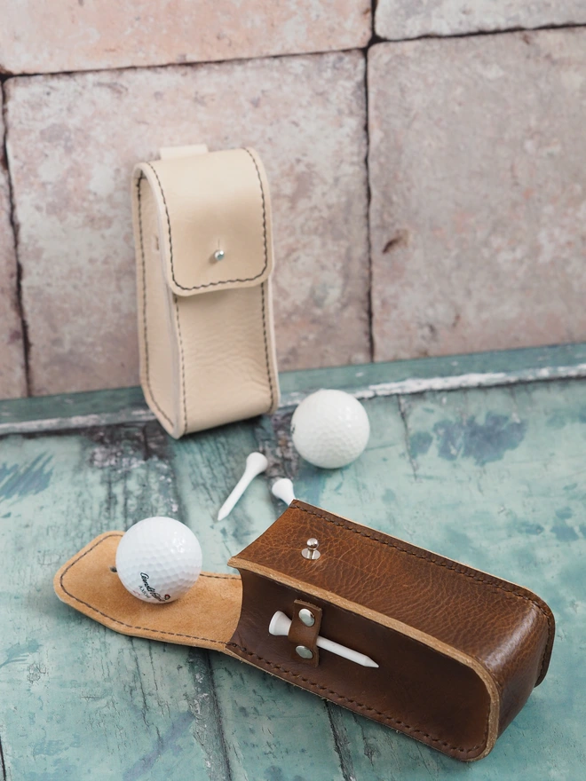 Handmade leather golf ball and tee carry case. Two leather pouches, one in natural cream leather, the other hand dyed a tan brown colour. Showing golf balls and tee accessories that they can carry.