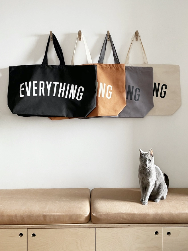Four colours of the Everything oversized tote bag hanging on coat hooks, a cat sits on a bench below