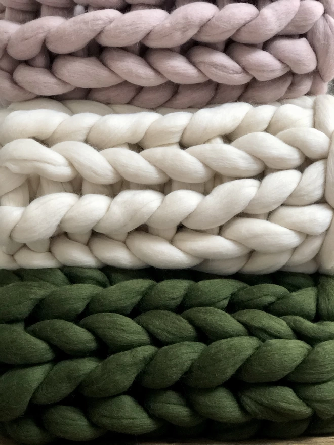 A close up of a stack of giant knitted blankets in mink pink, oyster white willow green