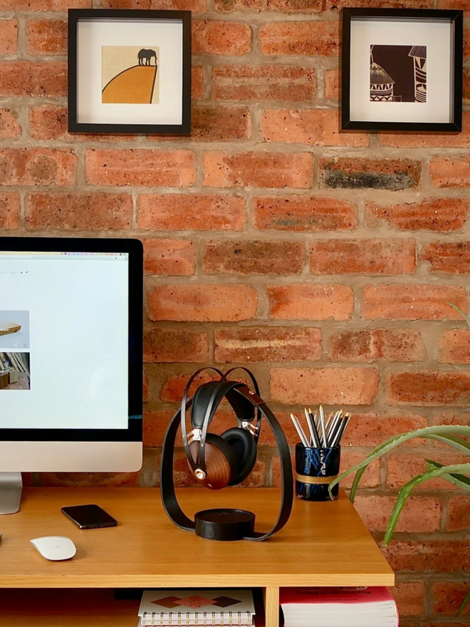 The Band Wooden Headphone Stand. A wooden headphone stand, made of one bent piece of wood, sat on a desk and in use with a pair of headphones. The stand is Ebonised (Black) with Brown leather.