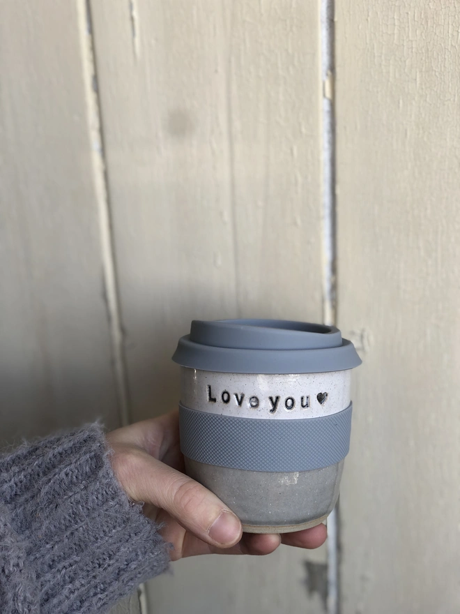 Personalised Short Ceramic Travel Mug, a blue ceramic travel mug sitting being held by a person in front of a wooden background. It's personalisation reads: ' Love you'. 