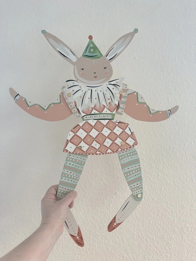 Large Hope Hare Wall Puppet. a hand holds the large hope hare wall puppet up against a plain wall, showing its jointed wooden limbs painted in soft pink, green and white tones. the hare wears a harlequin-style outfit with a diamond-patterned skirt, striped tights and a ruffled white collar, topped with a small pointed party hat. brass fasteners at the shoulders, hips and knees allow the limbs to move, giving the puppet a playful, articulated character.