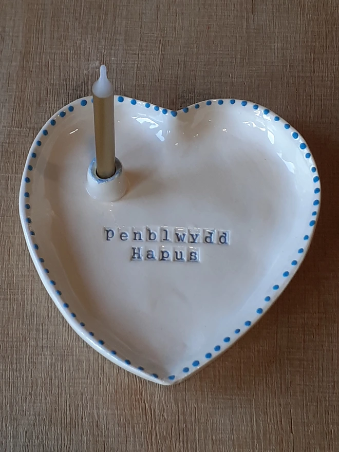 Handmade white heart-shaped ceramic birthday dish featuring the Welsh greeting “Penblwydd Hapus” in blue lettering, with blue dotted edging and a candle in the built-in holder.