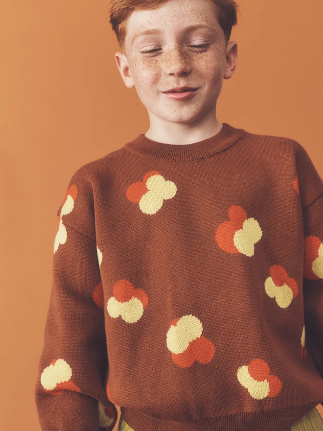 hild stands facing camera against a burnt orange backdrop, wearing a Studio Koter chestnut jacquard jumper with yellow and orange floral motif and chartreuse wide-leg corduroy trousers.