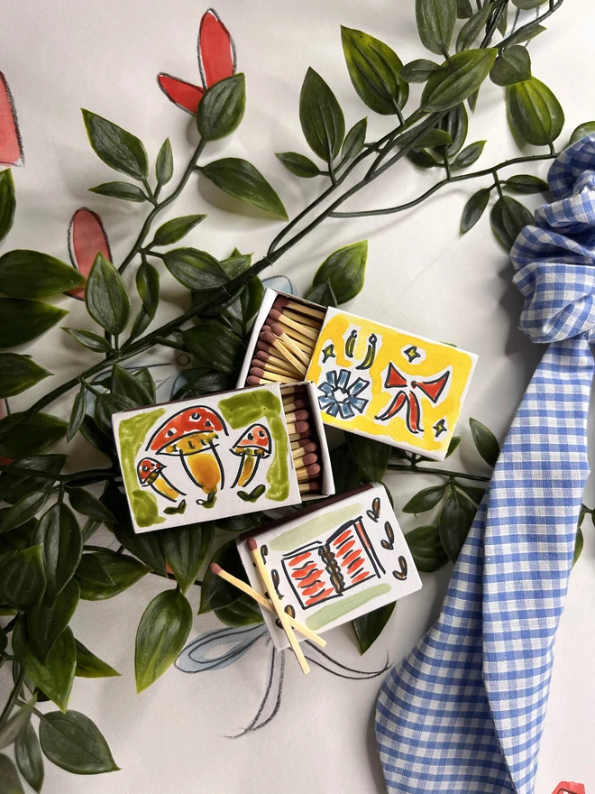 A whimsical arrangement featuring hand-painted matchboxes with vibrant designs, including mushrooms, abstract patterns, and a book.