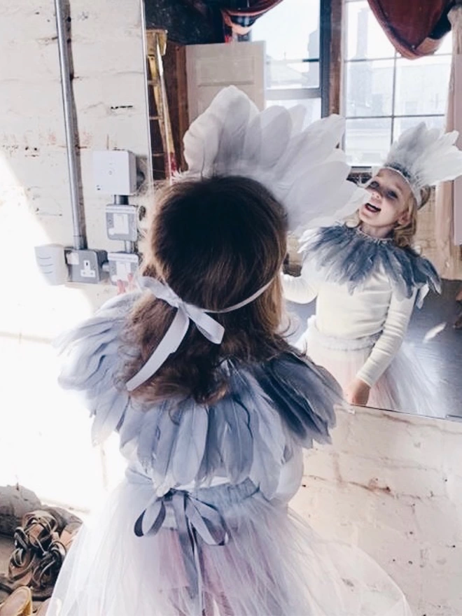 Smiling Girl looking in the mirror, wearing a grey feather headdress, grey feather cape and grey romantic tutu 